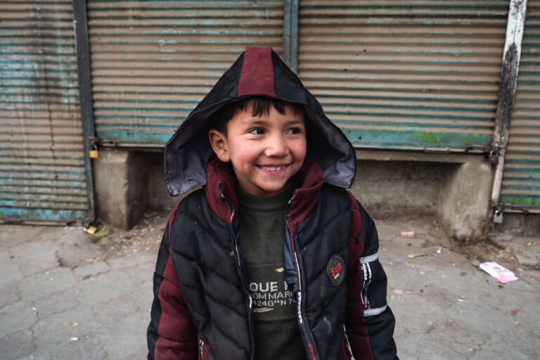 Smiling Afghan child in warm clothes standing in front of a closed shop door in Kabul, Afghanistan.