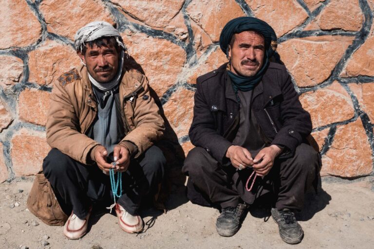 Two Afghan men sitting against a stone wall, smiling warmly in the chilly outdoors of Bamyan, Afghanistan.