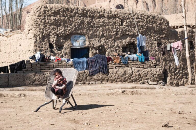 A girl sitting in a wheelbarrow in front of mud-brick houses, surrounded by mountainous terrain in Bamyan, Afghanistan.