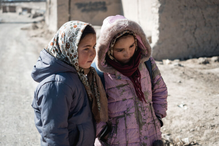 Two Afghan girls in winter clothes standing together on a dirt path with a village backdrop in Bamyan, Afghanistan.