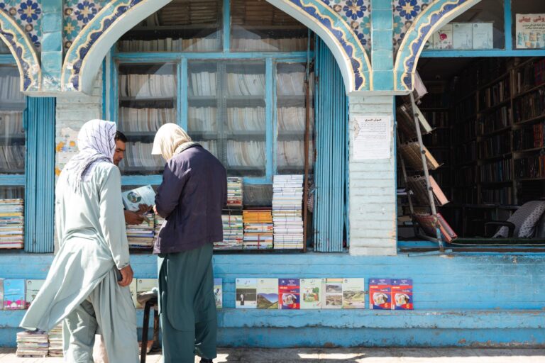 Three men buying books in front of a blue library surrounded by traditional Afghan architecture in Mazar-i-Sharif, Afghanistan.