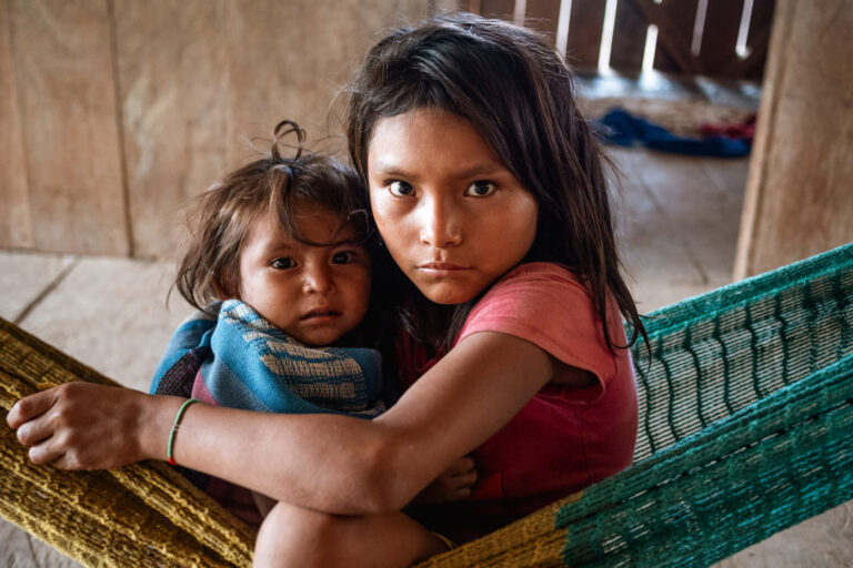 Young Achuar girl in a woven hammock, holding her sibling, looking pensive.
