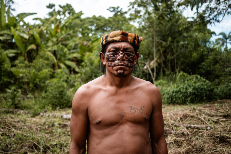 Shirtless Achuar elder smiling proudly in a lush Amazonian environment.