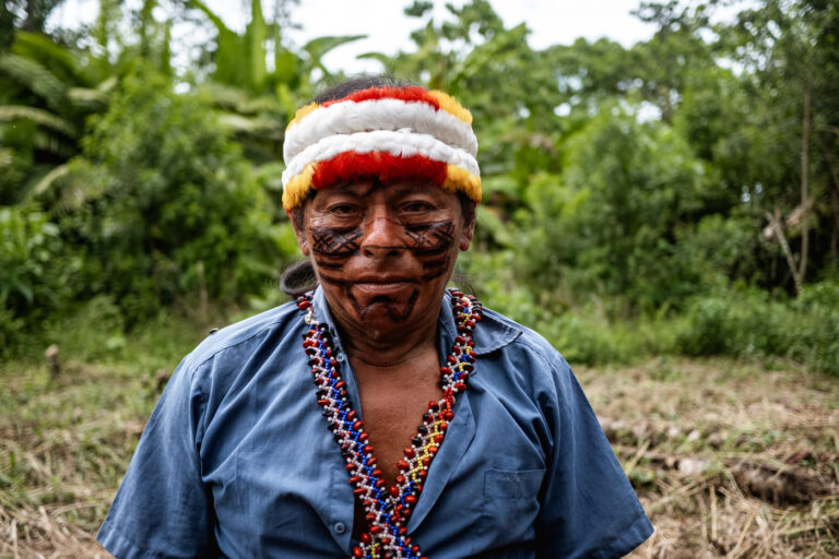 Achuar elder with red headband and beaded necklace, standing in jungle clearing.