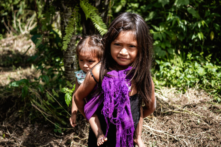 Two Achuar girls in traditional dress, standing in dense green Amazon jungle