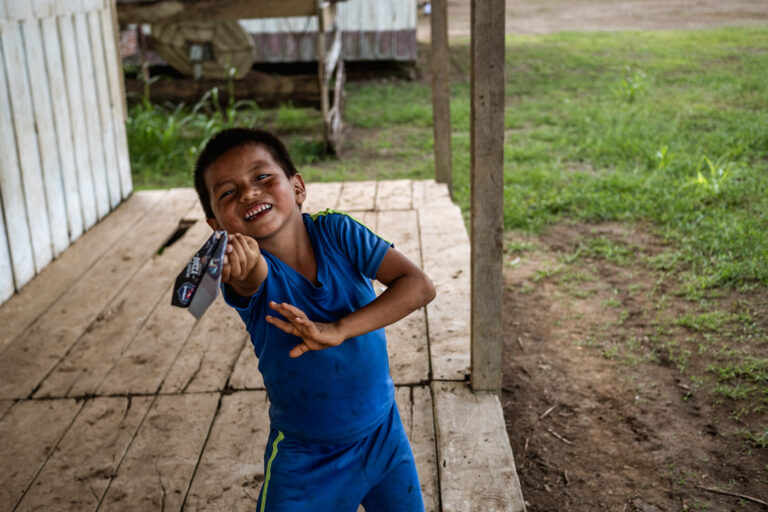 Young Achuar boy pointing, standing in front of a wooden Amazonian house.