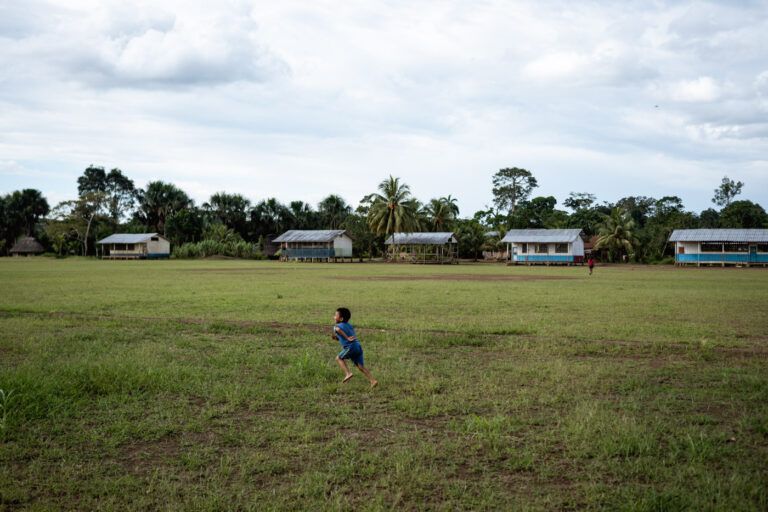 Achuar children running on grassy field, village houses in the background.