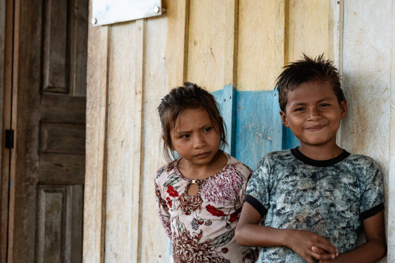 Two young Achuar children, smiling on wooden porch of their home.
