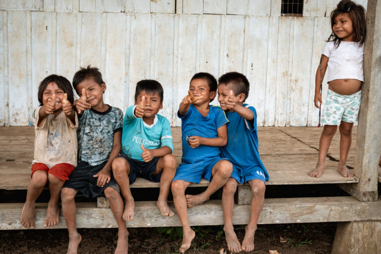 Laughter and joy: Achuar children posing together against wooden house wall.