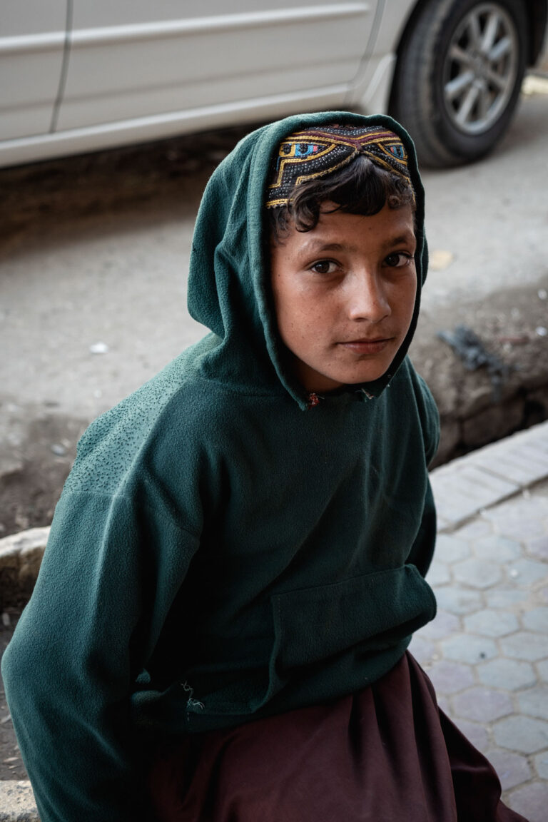 Afghan boy in a green hooded garment standing near a car, with a serious expression.