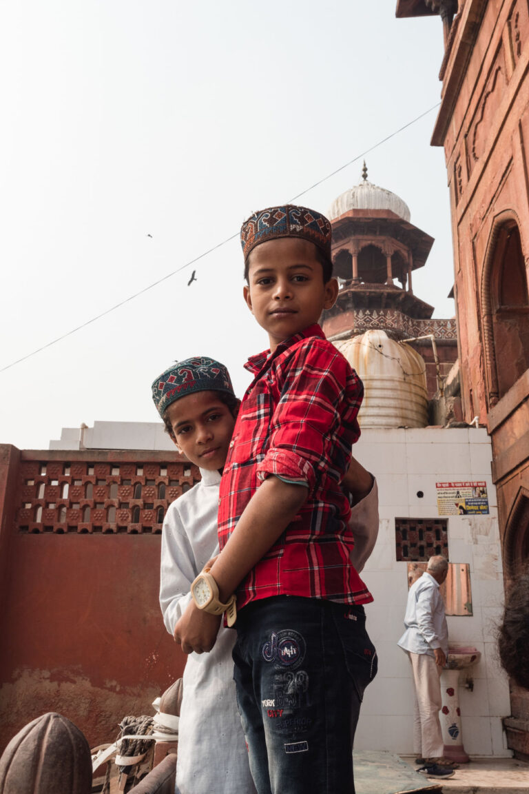 Two smiling children standing on a staircase, with colorful traditional Indian architecture in the background.