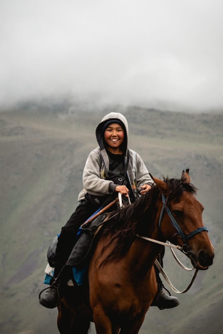 Kazakh man in traditional attire riding a horse across a vast, windswept mountain plateau.