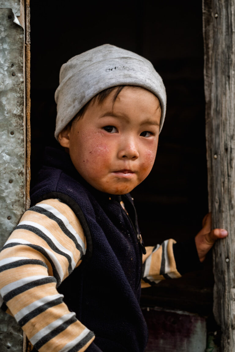 Kyrgyz boy peeking out from behind a wooden door with a curious and intense gaze.