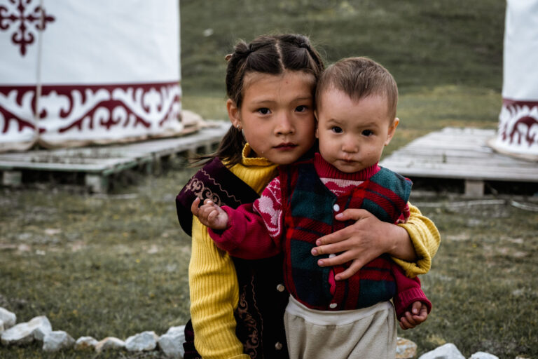 Two Kyrgyz siblings in colorful jackets sitting side by side, sharing a joyful moment.