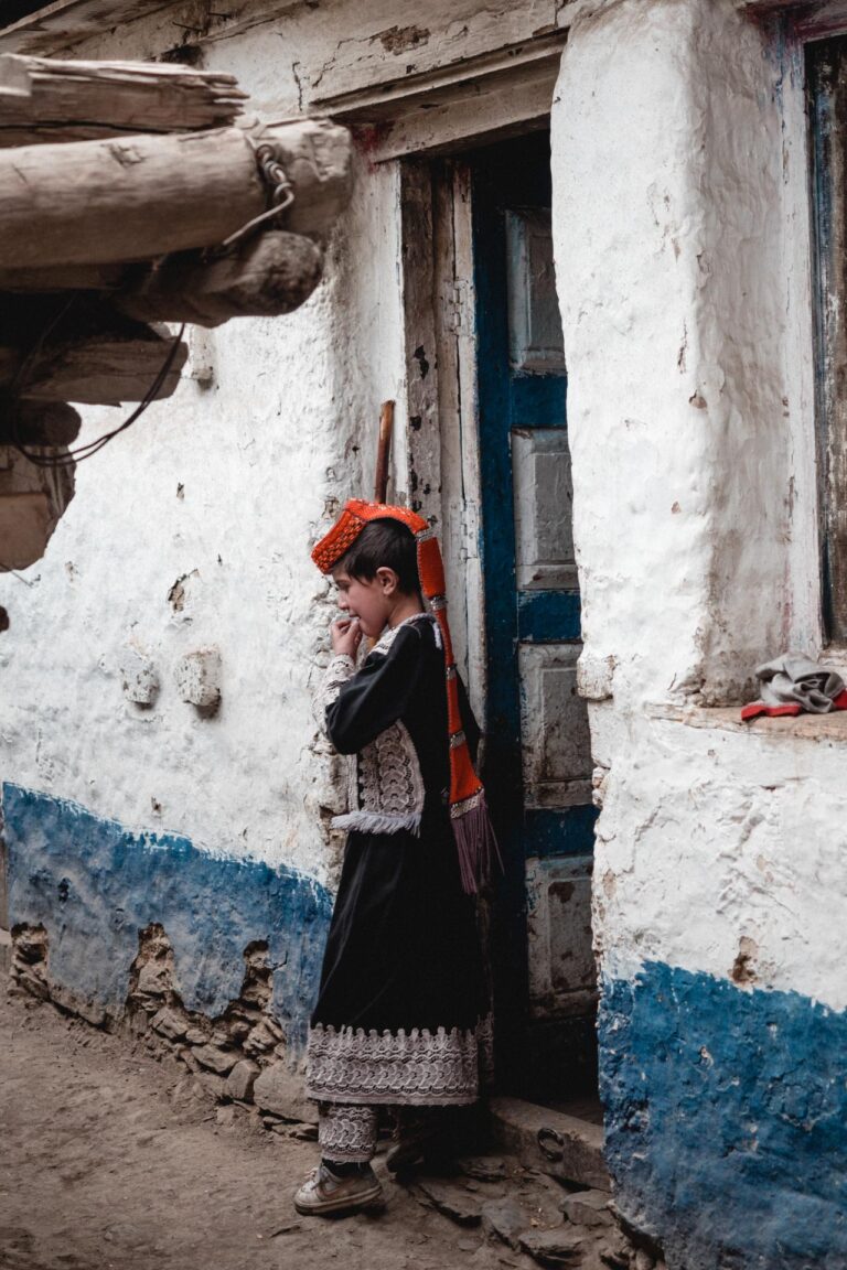 Kalash girl in a vibrant traditional headdress, standing in a mud-brick alley in Bumburet Valley.