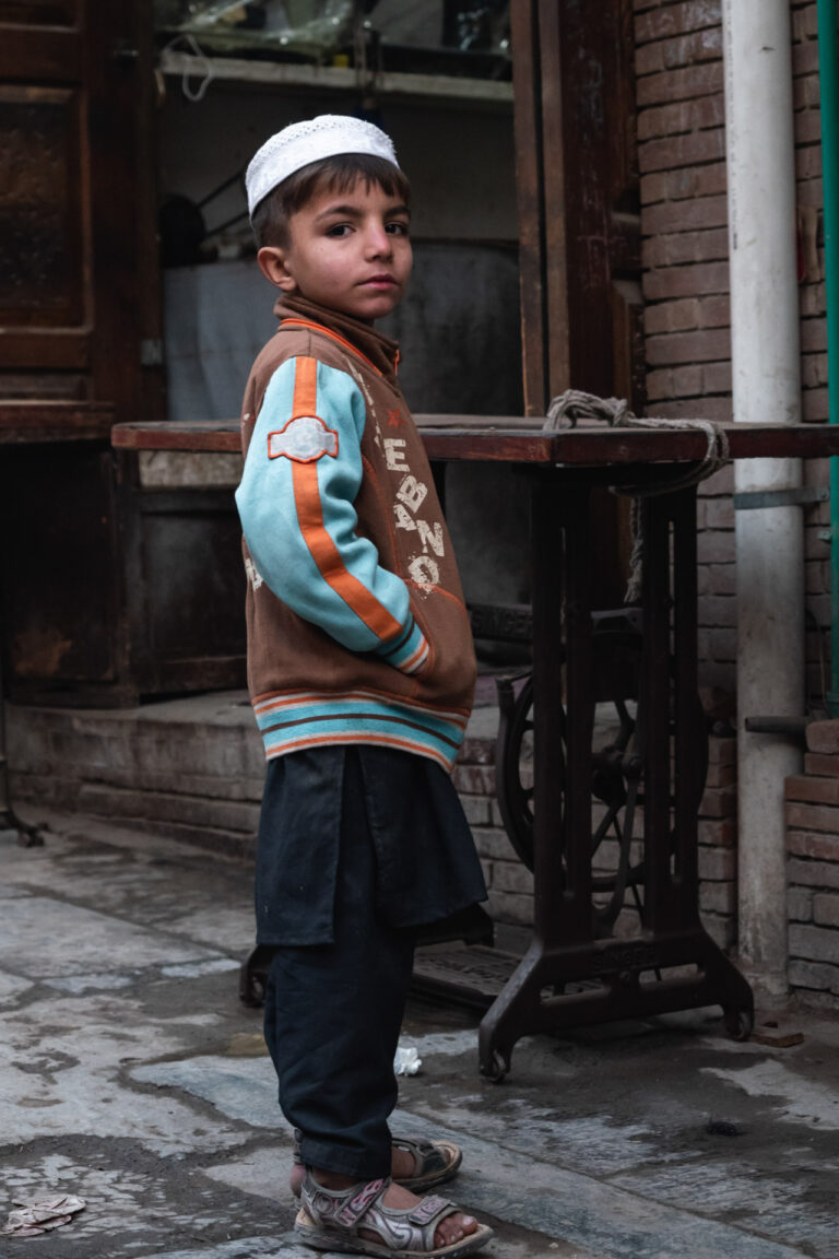 Young Pakistani boy wearing a cap and vest, standing confidently against a brick wall.