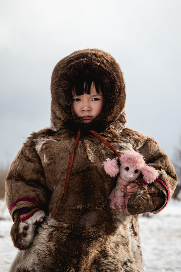 Nenet girl in fur coat holding plush toy, Siberian winter landscape.