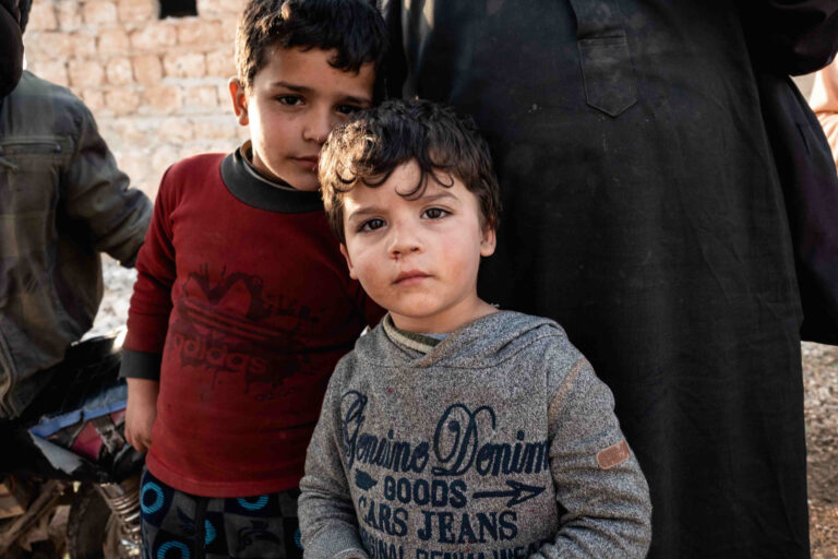 Two young boys stand together, one looking curious, the other slightly reserved, behind adults.
