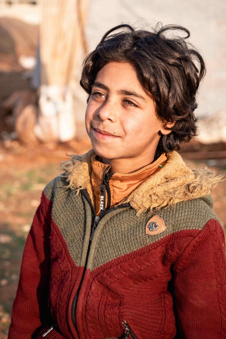 A smiling boy in a red and brown jacket stands in golden sunlight, eyes bright.