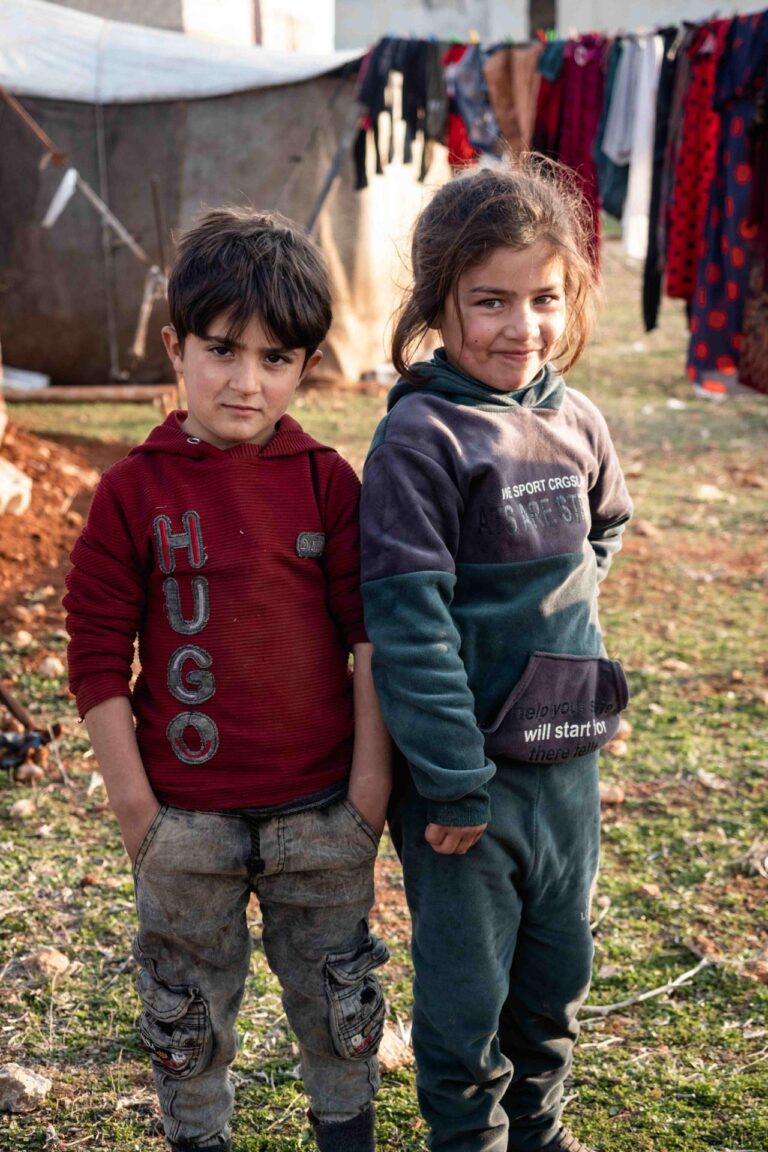 Two children stand near tents and hanging laundry, wearing warm clothes, gazing curiously at the camera.