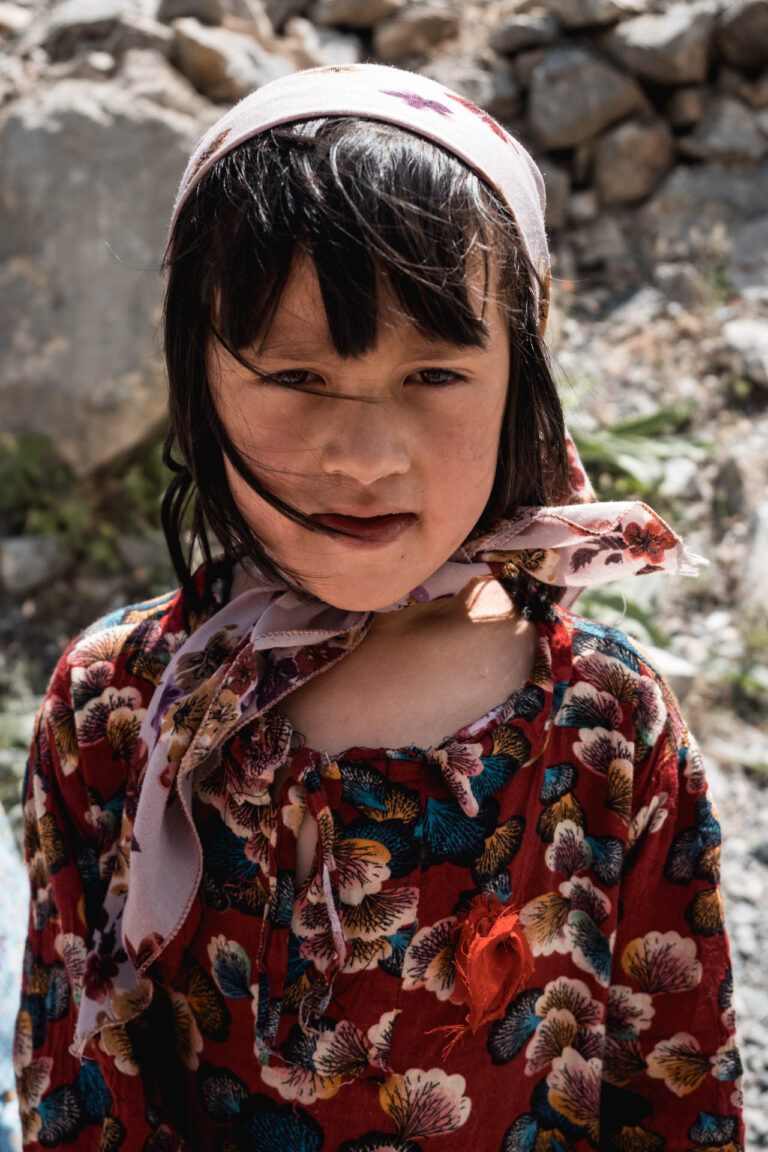 Smiling Tajik girl in colorful dress standing against a backdrop of rugged mountains.