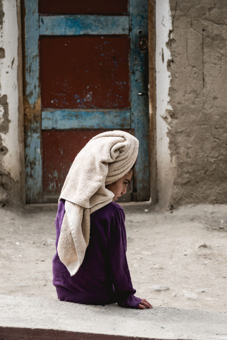 Tajik girl in traditional attire with her back to a mud-brick doorway, holding her headscarf.