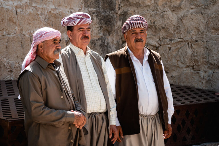 Elderly Yazidi men discussing traditions near Lalish's historic stone walls