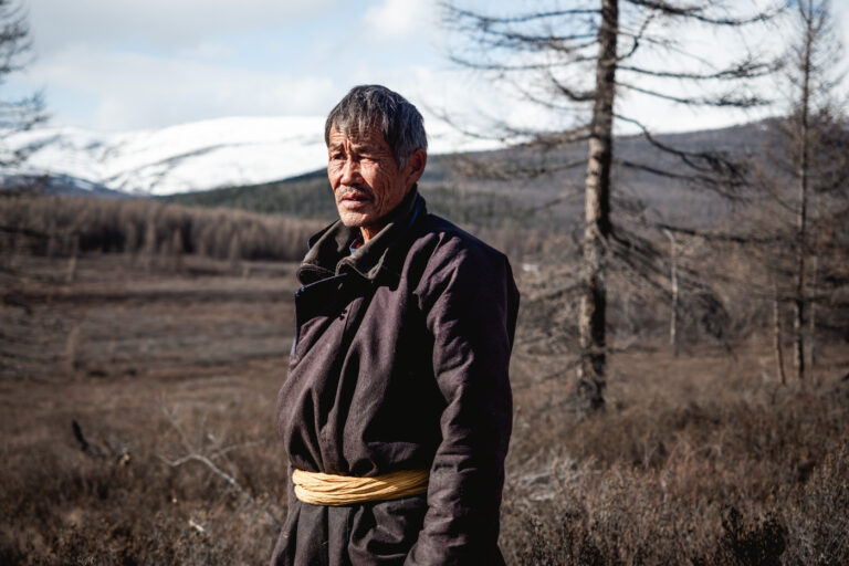 Tsaatan man standing outdoors, with forest and mountains in the background.