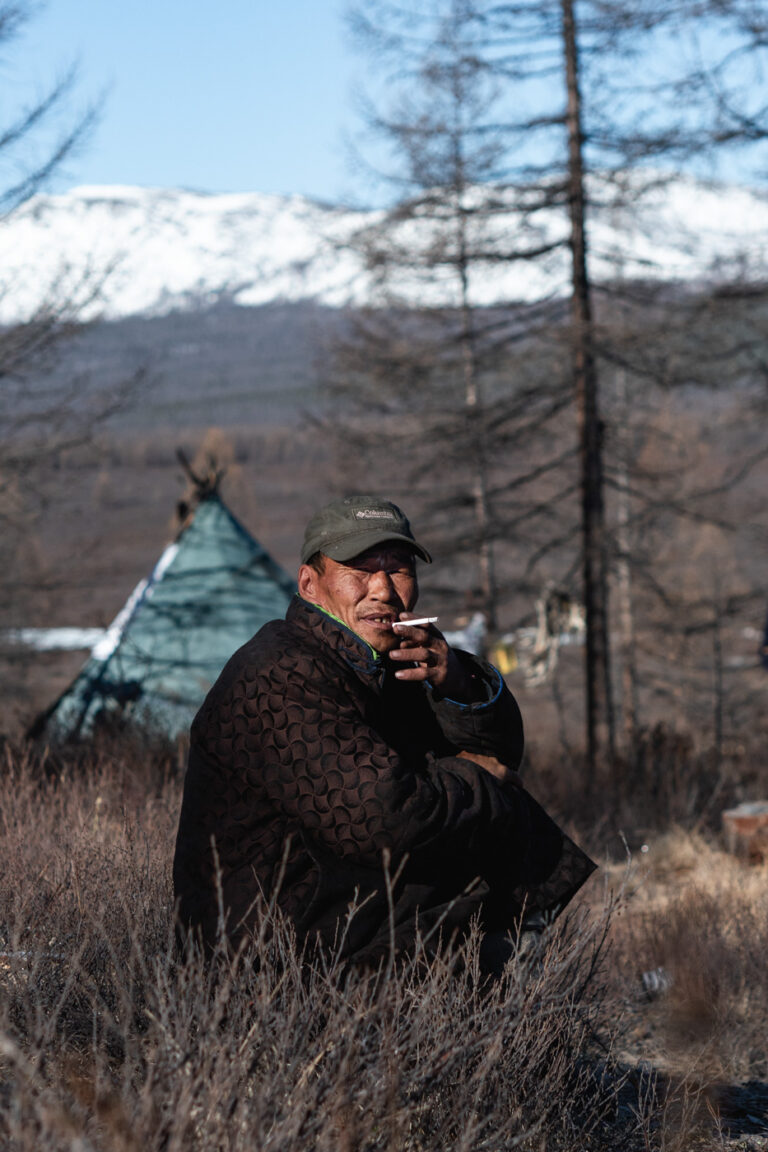 Tsaatan man riding reindeer between larch trees in the taiga.