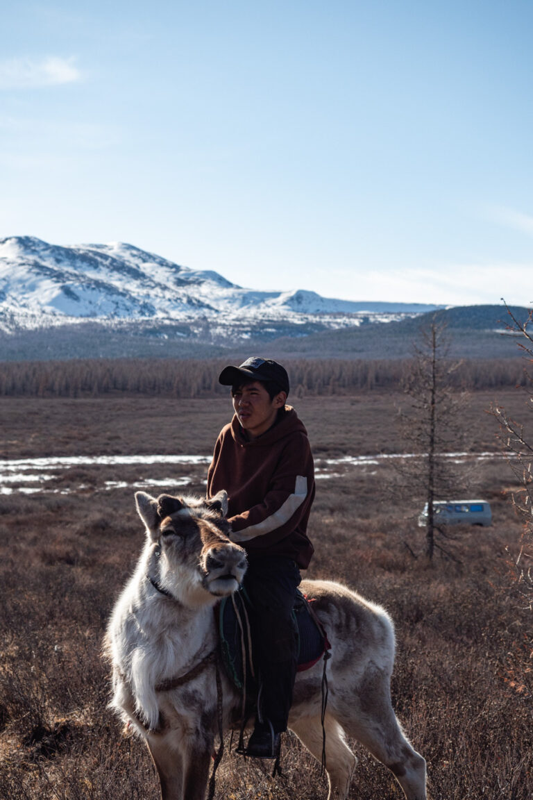 Tsaatan youth riding reindeer, distant mountain range behind.