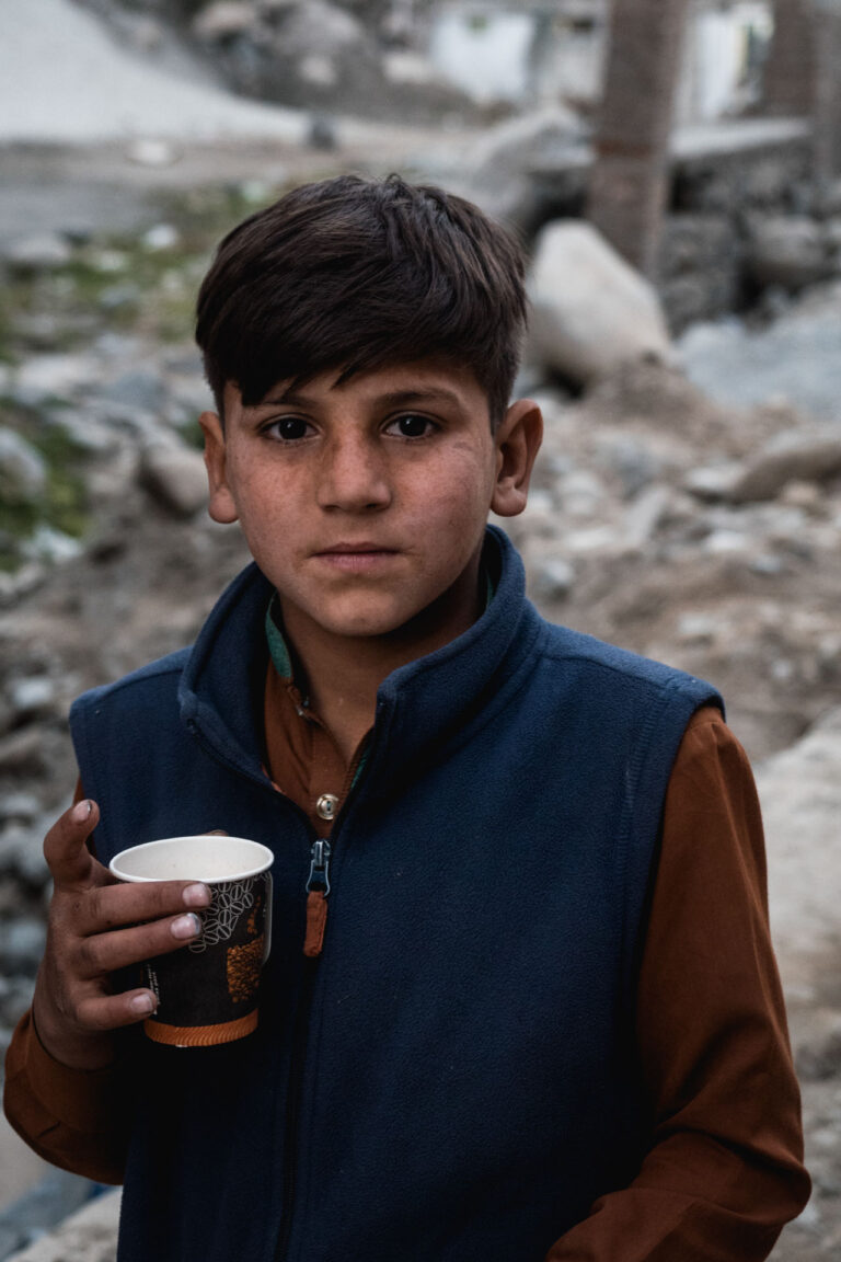 Young boy holding a steaming cup, standing in front of rustic wooden walls in Bahrein, Swat valley, Pakistan.