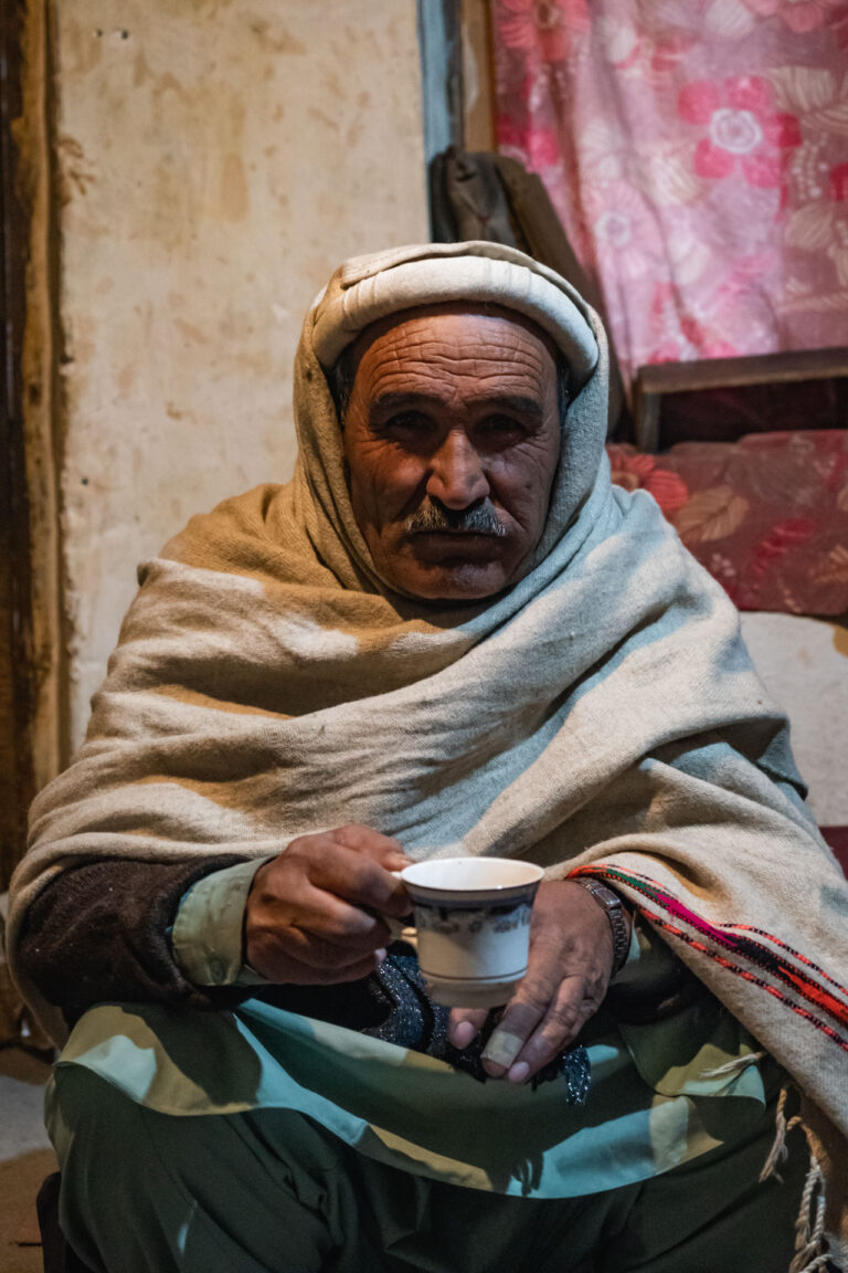 Elderly Chitrali man wrapped in traditional shawls sitting inside a warm space in Garam Chashma, Pakistan.