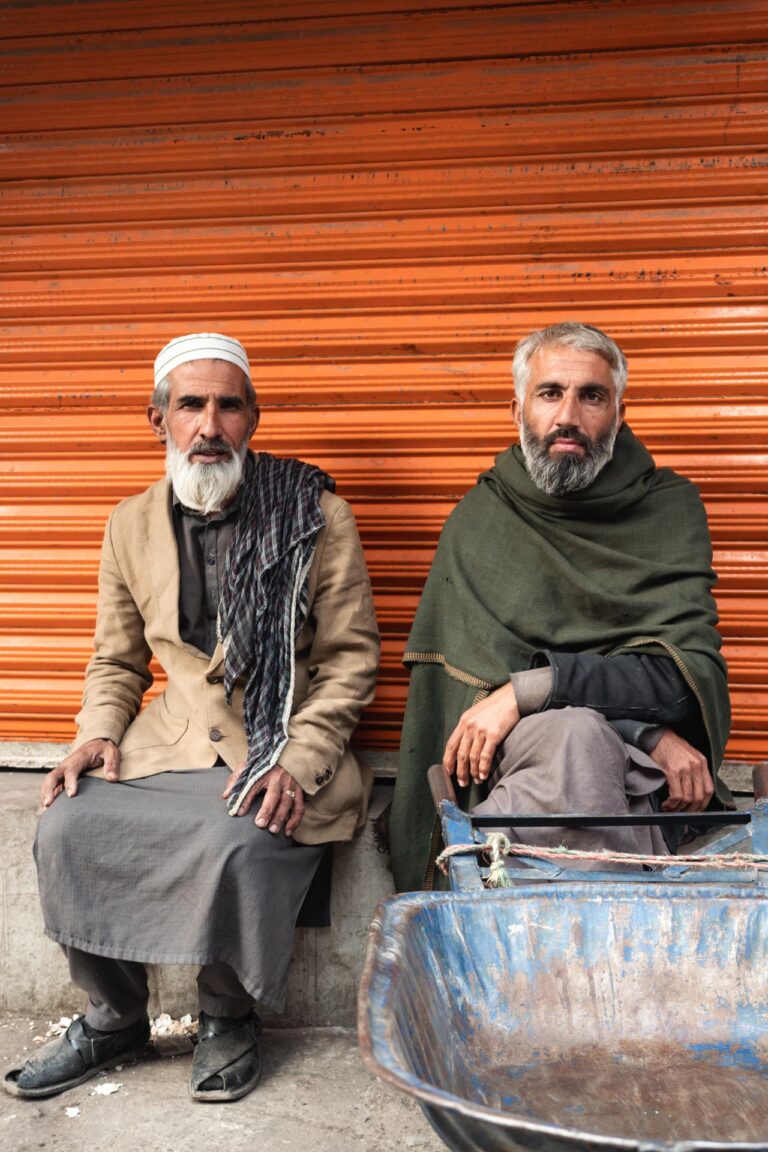 Two elderly men in traditional Pashtun attire standing together near a weathered wall in Peshawar, Pakistan.