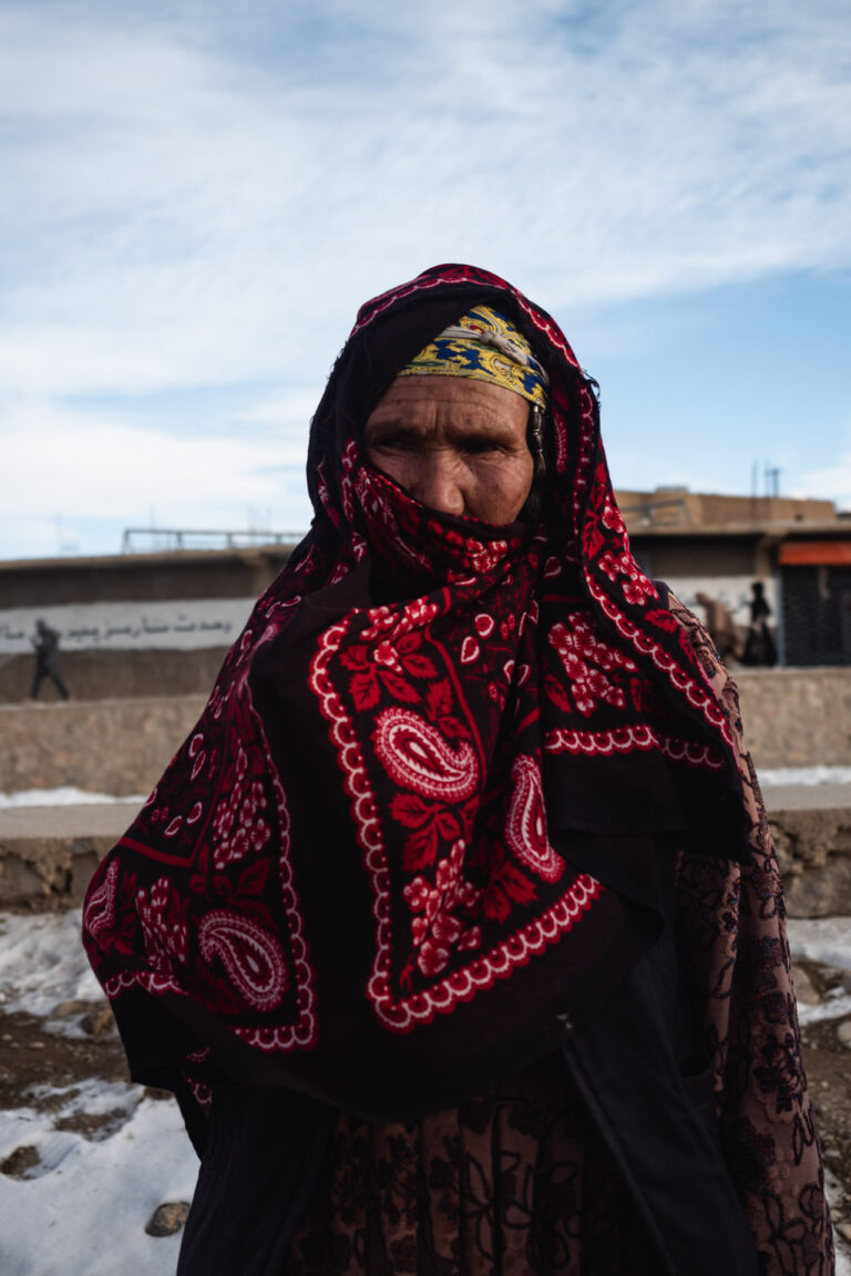 Erdely woman wearing a traditional embroidered shawl standing in front of the Bamyan cliffs in Afghanistan.