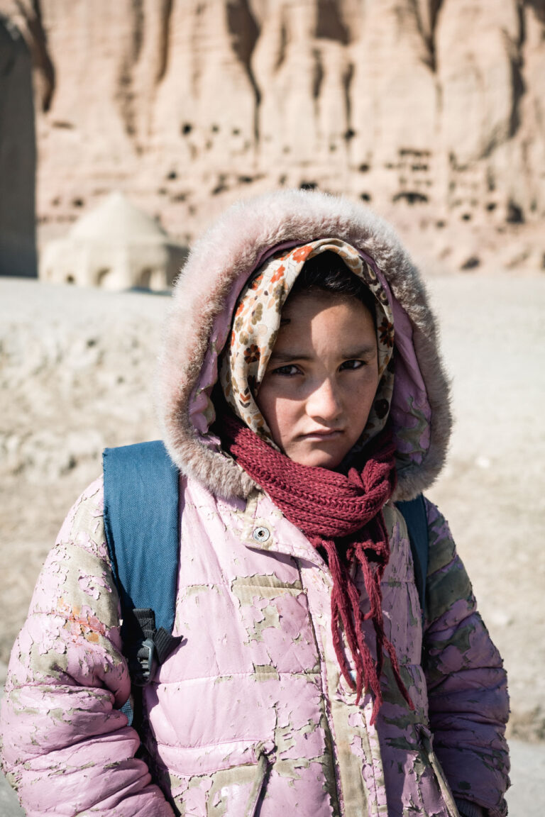 Girl standing in front of ancient Buddhist carvings in Bamyan, Afghanistan, wearing a pink scarf.