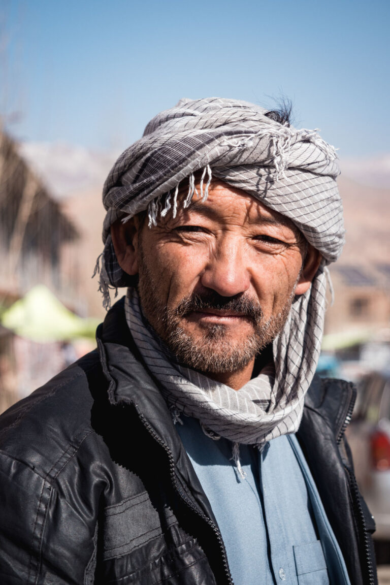 Smiling Afghan man wearing a white turban with snow-capped mountains in the background.