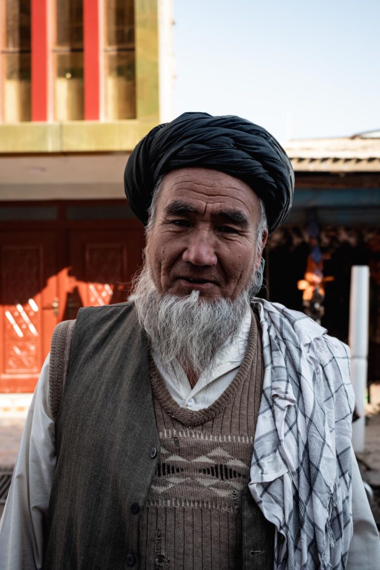 Elderly Afghan man with a long beard and woolen cap sitting indoors with a serious expression.