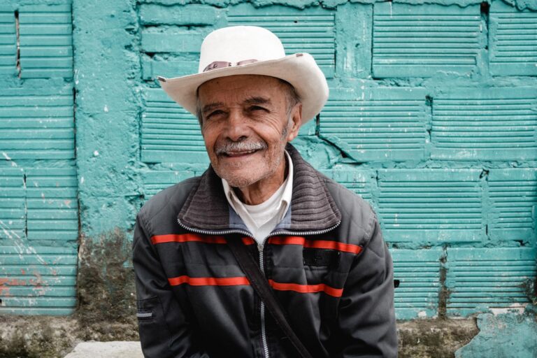 Elderly Colombian man wearing a hat and colorful shawl standing against a blue wall in Bogotá.
