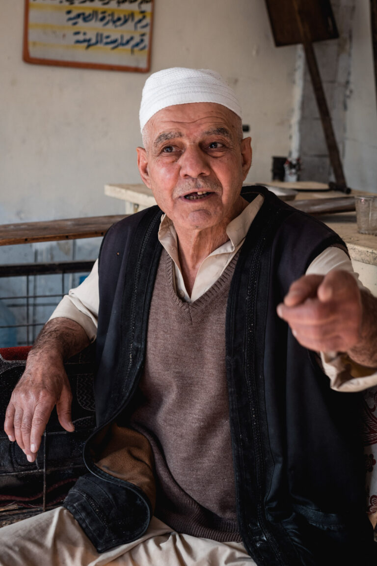 An elderly man sitting inside his sweet shop, inviting us to try his sweets, showcasing the hospitality found in Mosul, Iraq