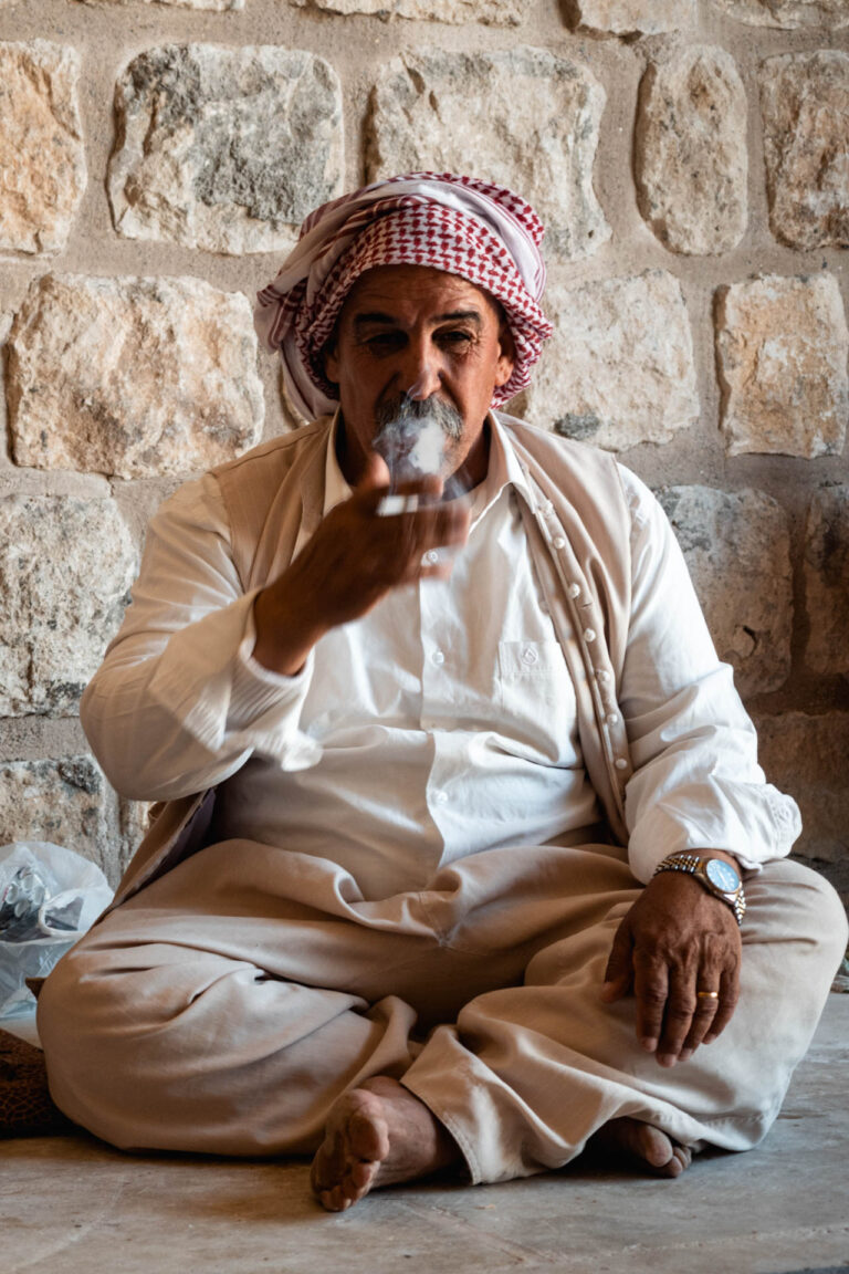 Elderly Yazidi man smoking a cigarette sitting against a stone wall in the sacred Lalish temple complex in Kurdistan.