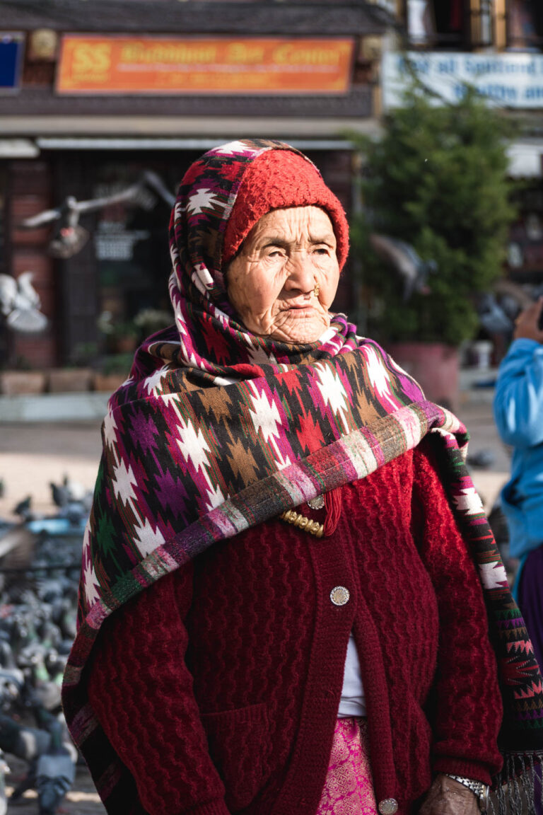 Elderly woman wearing a vibrant red shawl with prayer flags fluttering in the background near the Buddha Stupa.