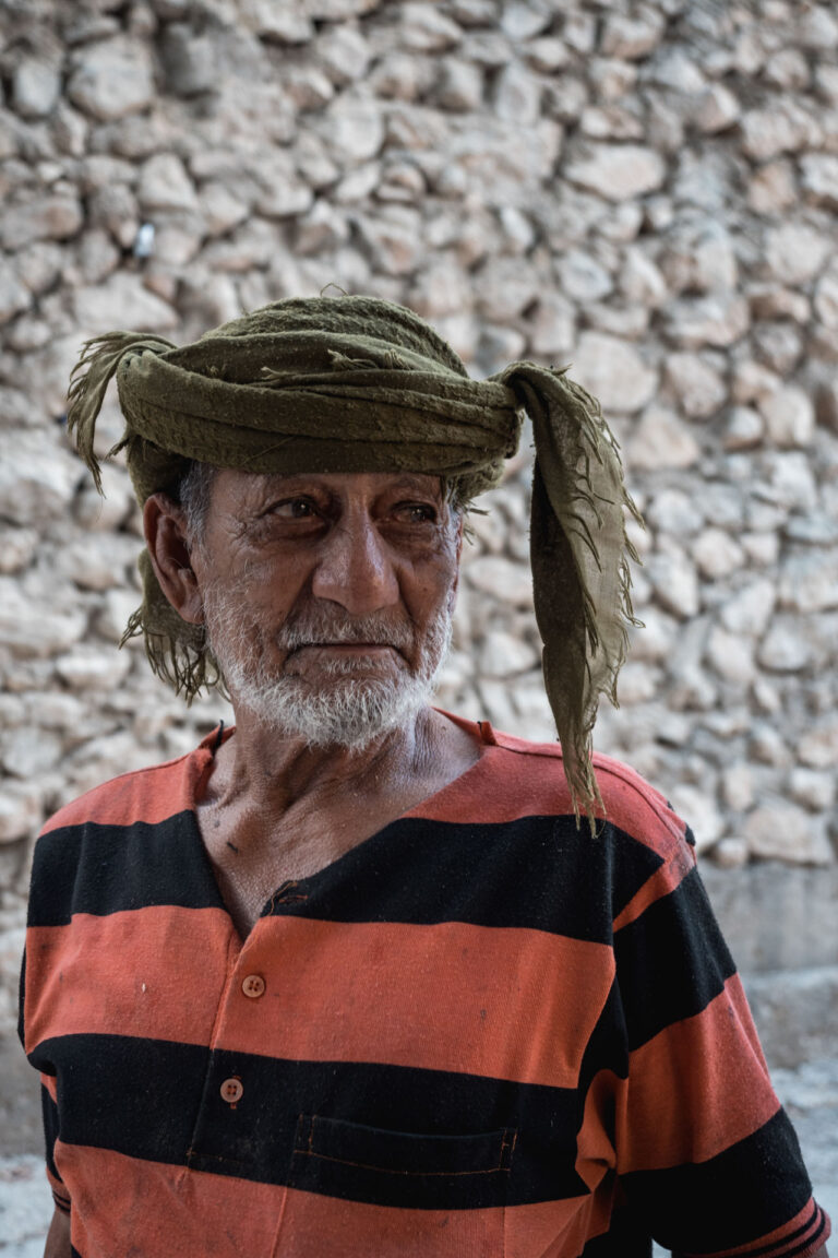 Portrait of an elderly man wearing a striped shirt standing against a stone wall in Wadi Tiwi, Oman.