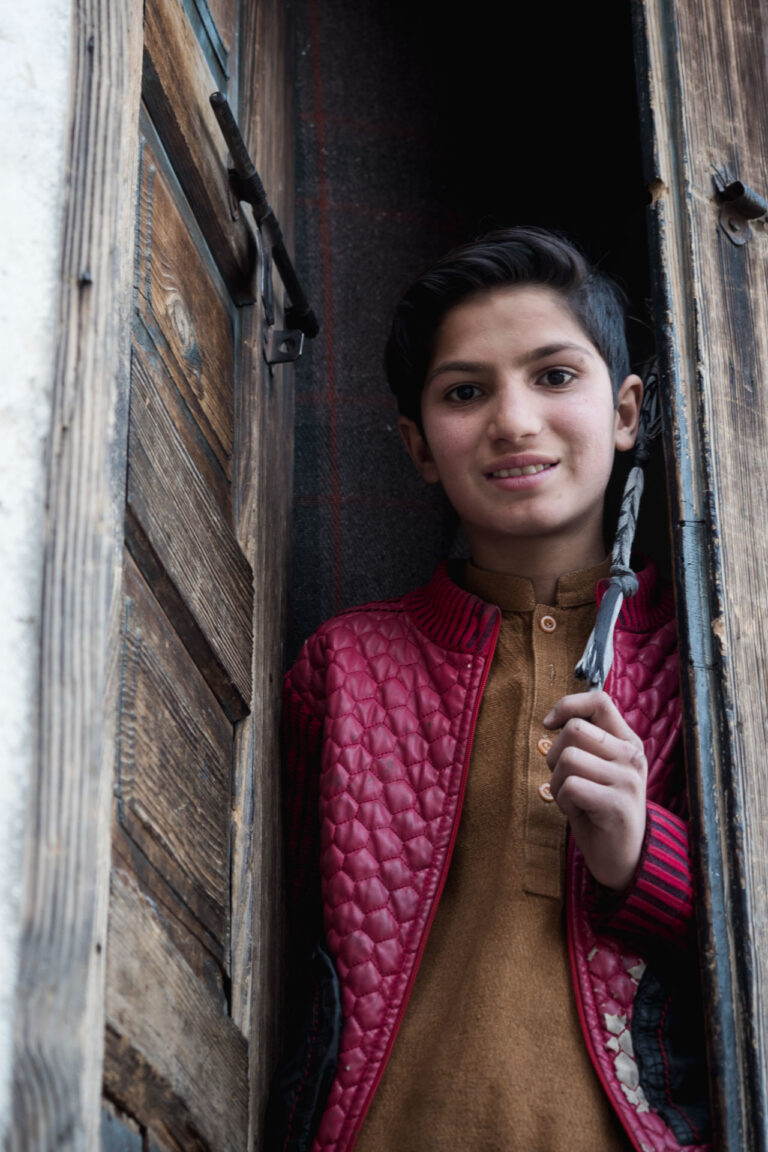 Young Pakistani boy in a red shawl standing outside a wooden doorway in Madyan, Swat Valley.