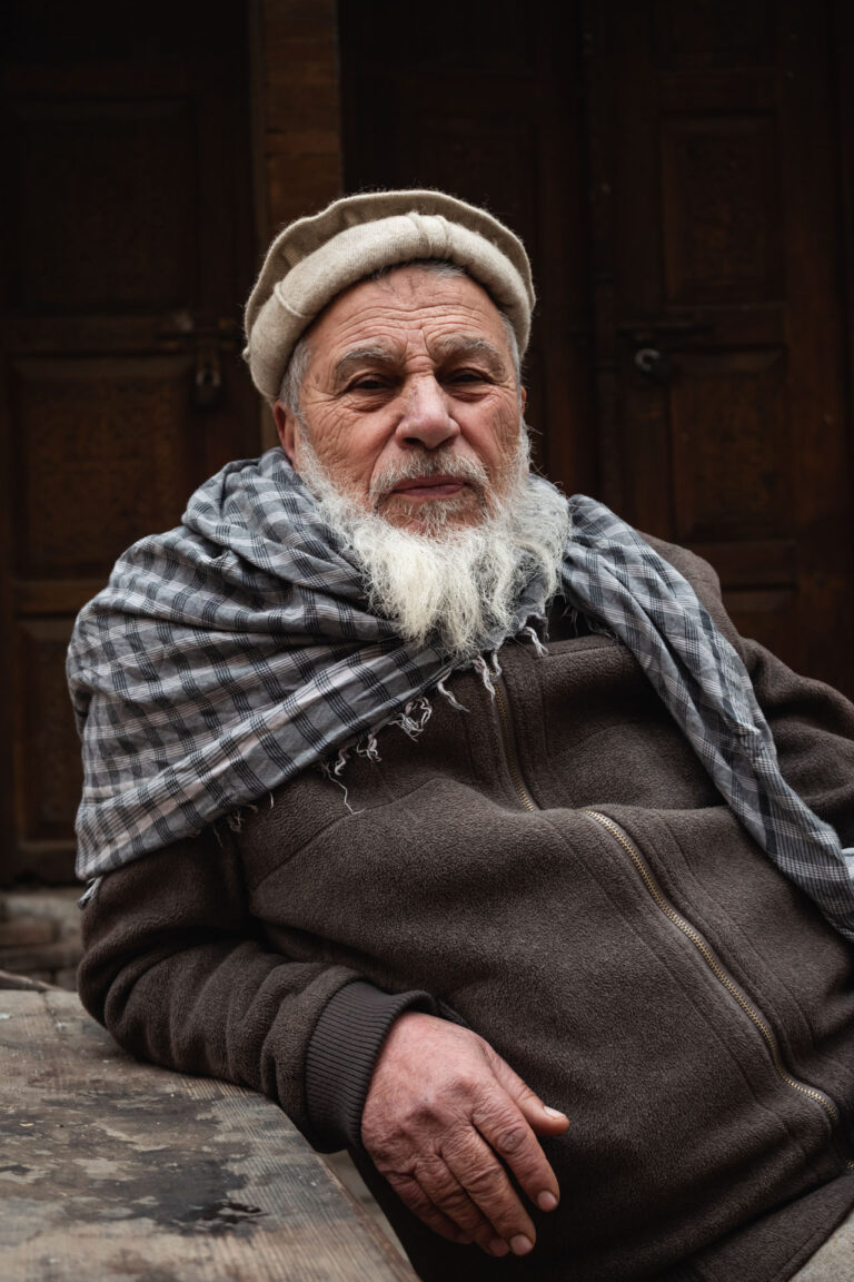 Elderly man sitting on a wooden stool wearing a thick woolen shawl in Peshawar.