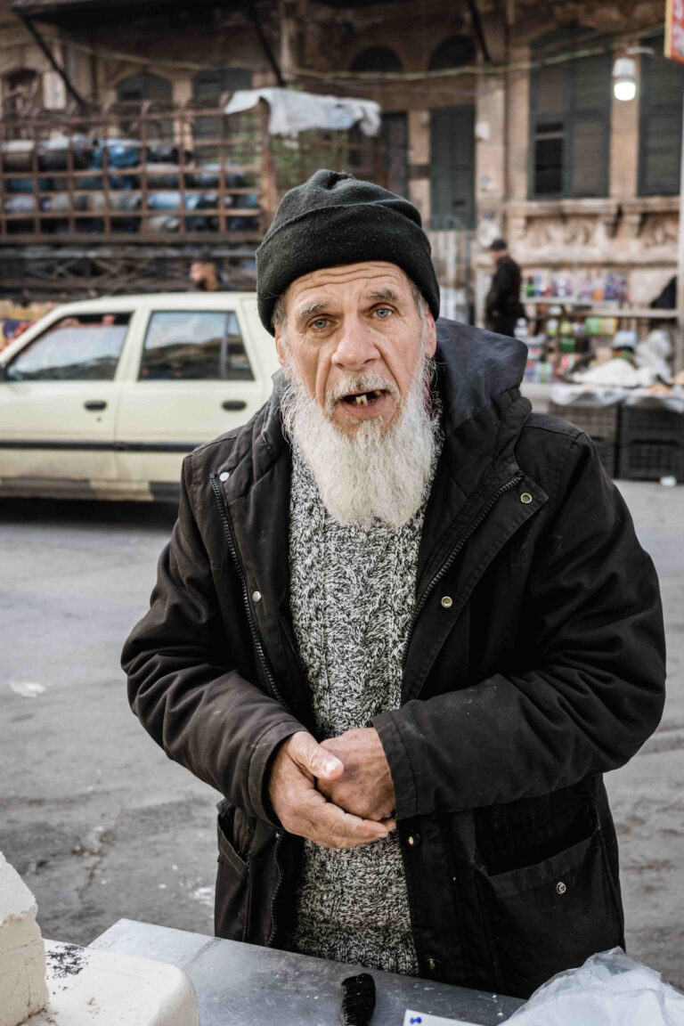 Elderly man with a long white beard, wearing a black beanie and winter coat, standing in a bustling marketplace in Aleppo, Syria.
