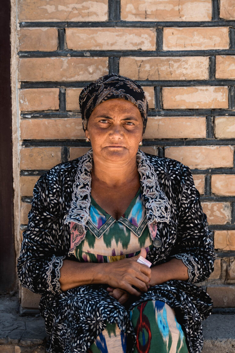 Elderly Uzbek woman sitting against a brick wall in Bukhara, wearing a colorful headscarf.