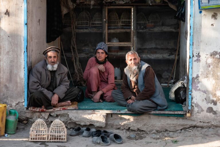 Afghan men sitting outdoors in front of their bird shops, looking directly at the camera, in Kunduz