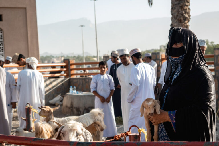 A group of Omani men dressed in white dishdashas, with a Bedouin woman in the foreground wearing her traditional mask, at the bustling Nizwa goat market