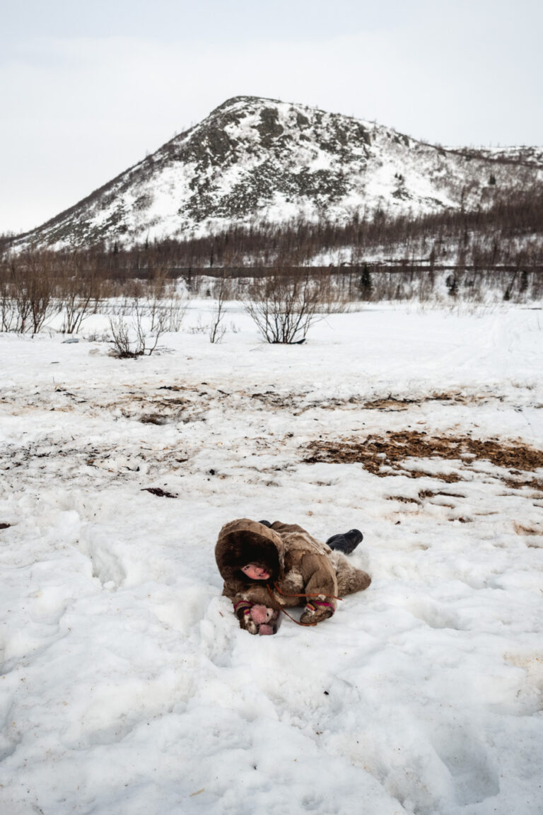Child curled up in fur blanket on snowy ground.
