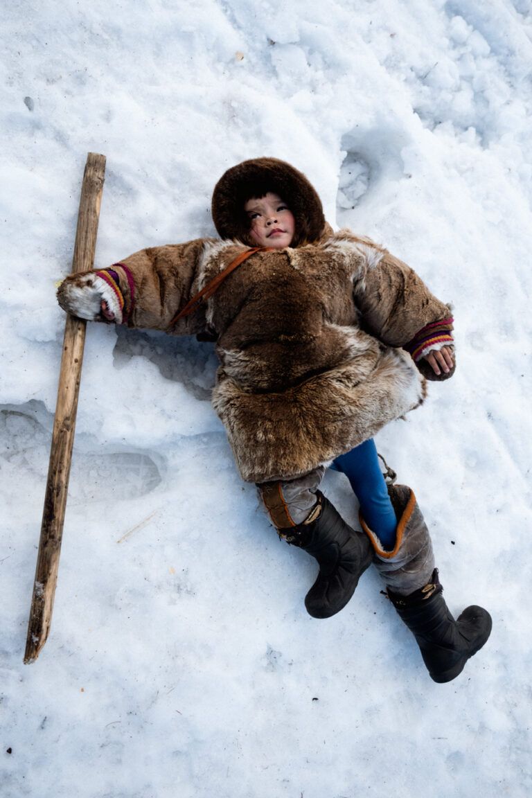 Nenets girl sitting alone on snow, wrapped in heavy fur coat.